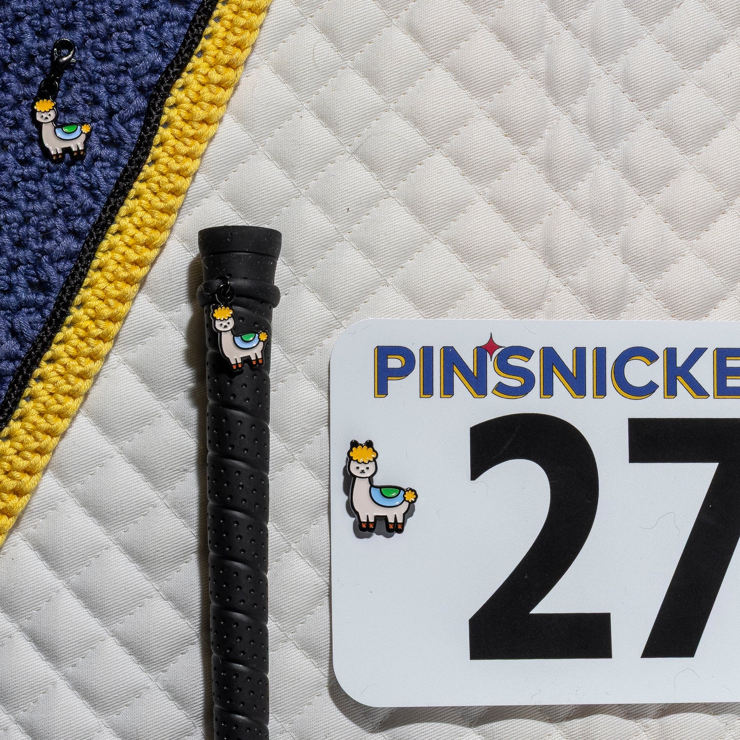 A saddle pad, bonnet and whip decorated with matching Drama llama Pinsnickety horse show number pins, a bridle charm and a crop charm.