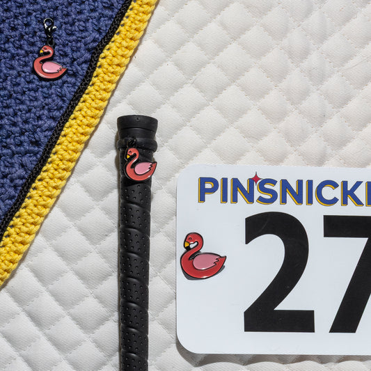 A saddle pad, bonnet and whip decorated with matching Flamingo Pinsnickety horse show number pins, a bridle charm and a crop charm.