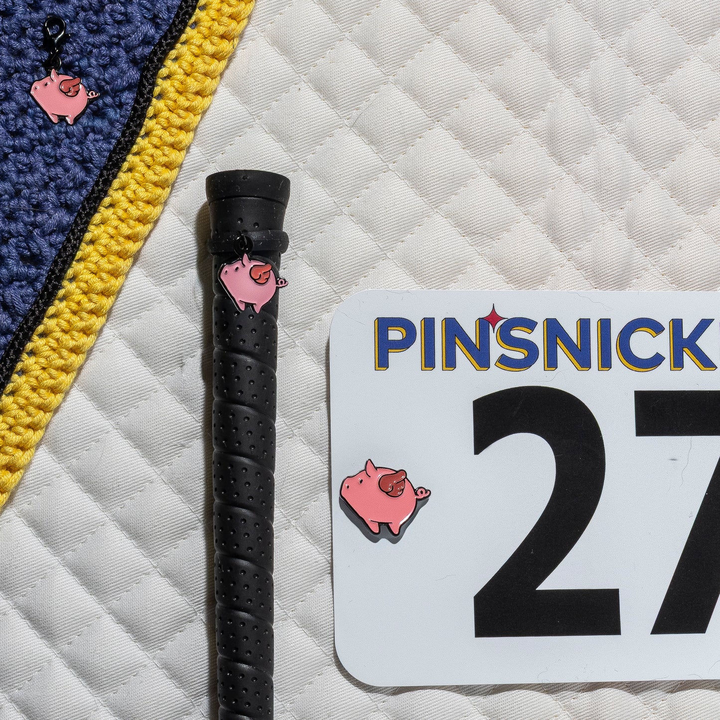 A saddle pad, bonnet and whip decorated with matching Flying Pig Pinsnickety horse show number pins, a bridle charm and a crop charm.