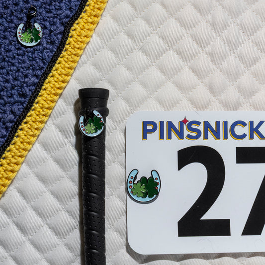 A saddle pad, bonnet and whip decorated with matching Hoeseshoe Pinsnickety horse show number pins, a bridle charm and a crop charm.
