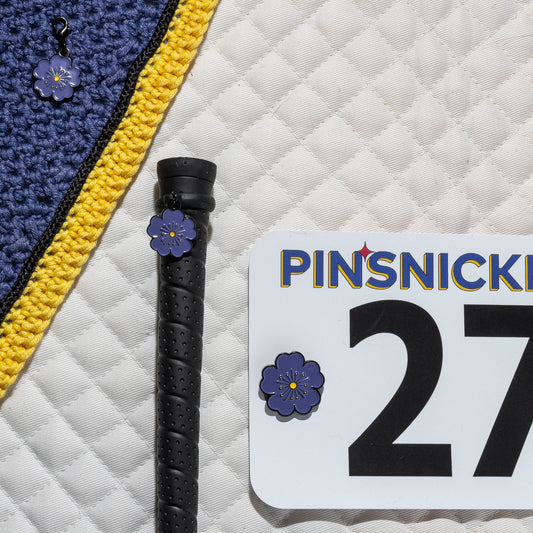 A saddle pad background showing a matching pinsnickety purple poppy number pin on a show number, a crop charm on a whip handle, and a bridle charm on a purple bonnet.
