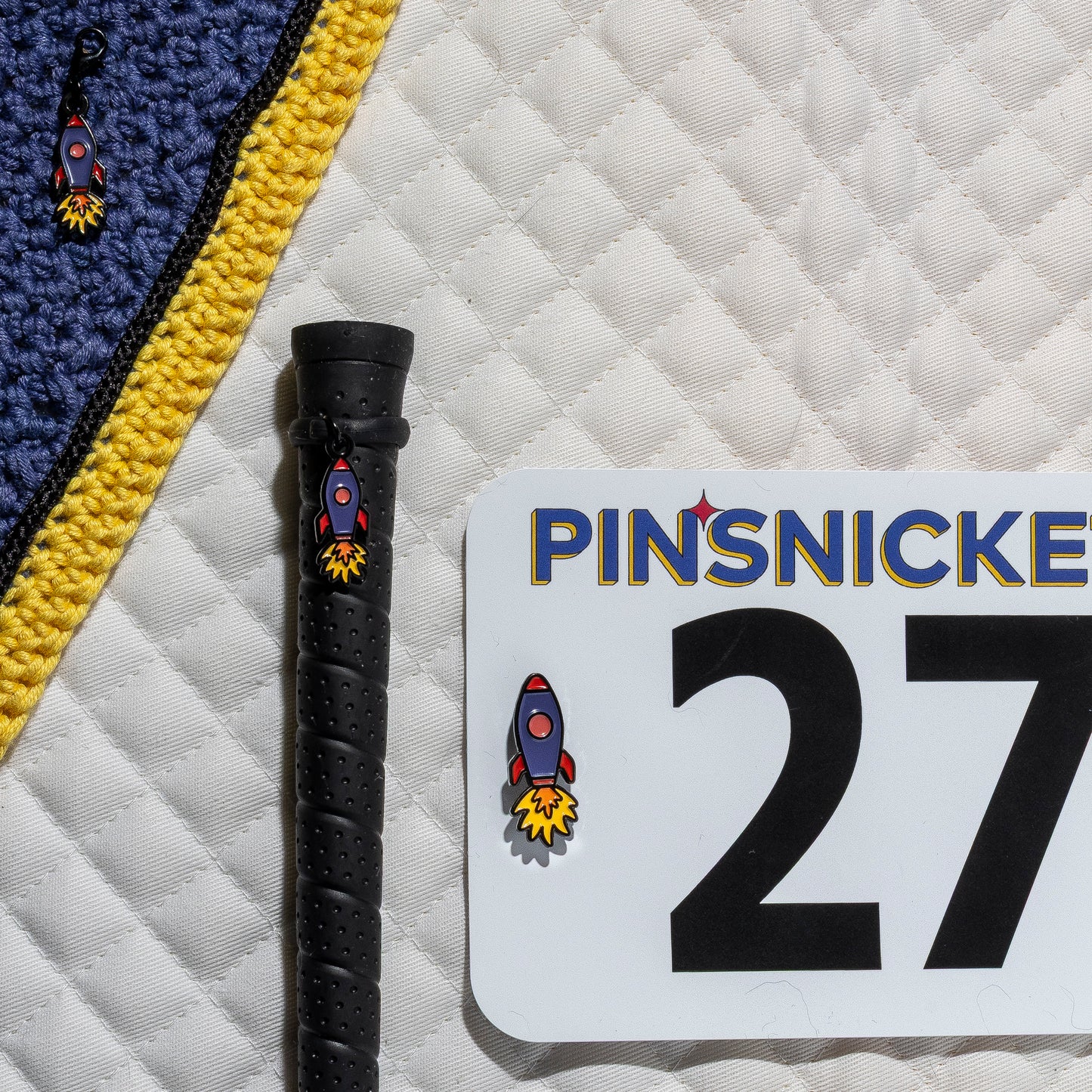 A saddle pad, bonnet and whip decorated with matching Rocket Ship Pinsnickety horse show number pins, a bridle charm and a crop charm.