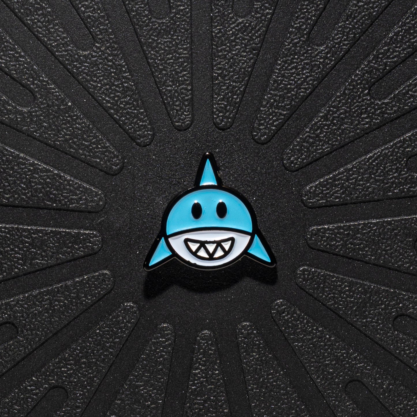 A single Pinsnickety horse show number pin shaped like a shark with a light blue body, black outline, and white teeth, centered on a textured black background with radiating grooves.