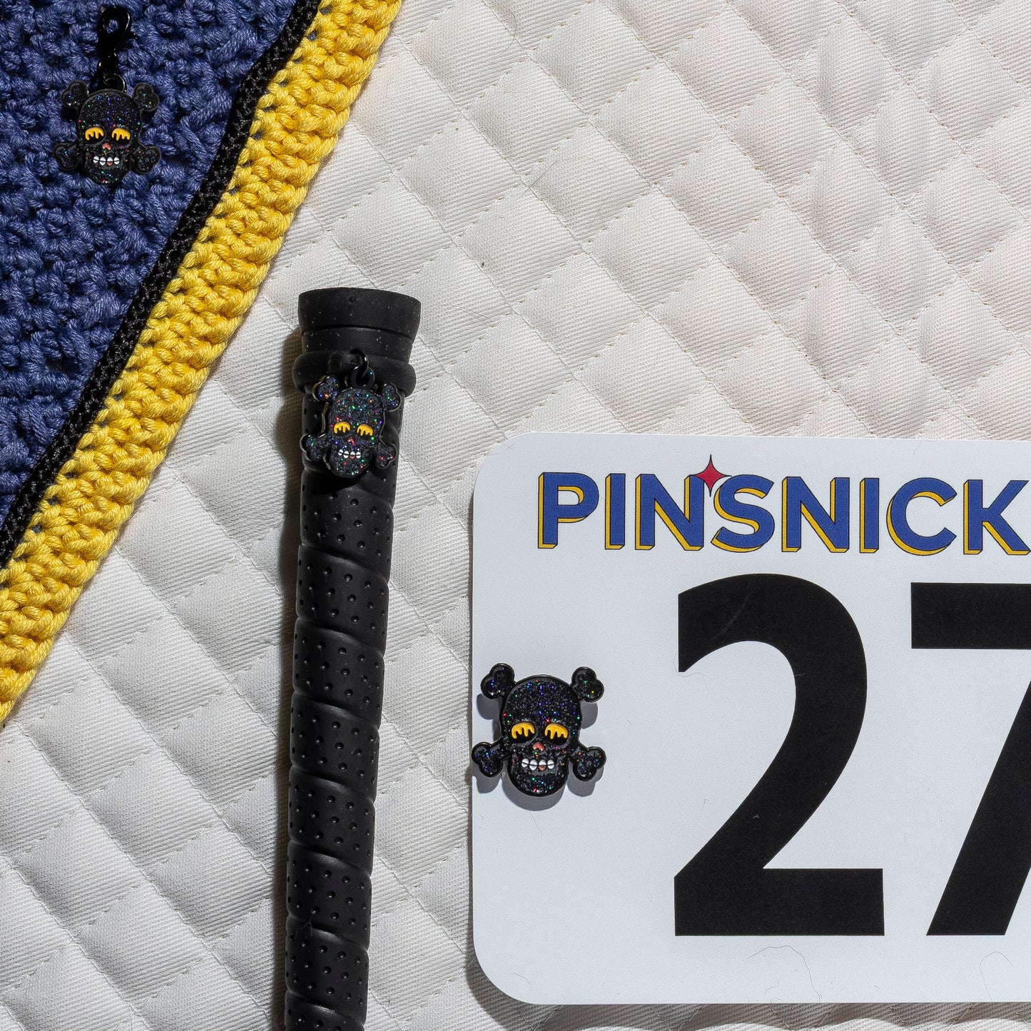 A saddle pad, bonnet and whip decorated with matching Skull Pinsnickety horse show number pins, a bridle charm and a crop charm.