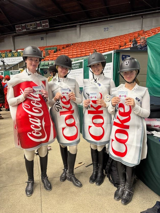 four young horseback riders in diet coke halloween costumes hold up pinsnickety horse show number pins in front of orange stands in an arena.