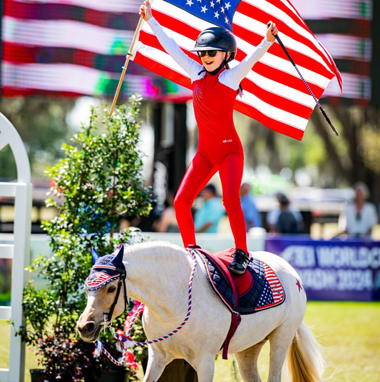 emma crawford standing on her pony Gabby and holding an american flag. gabby's bonnet and saddle pad are decorated with pinsnickety stars and stripes horse show number pins