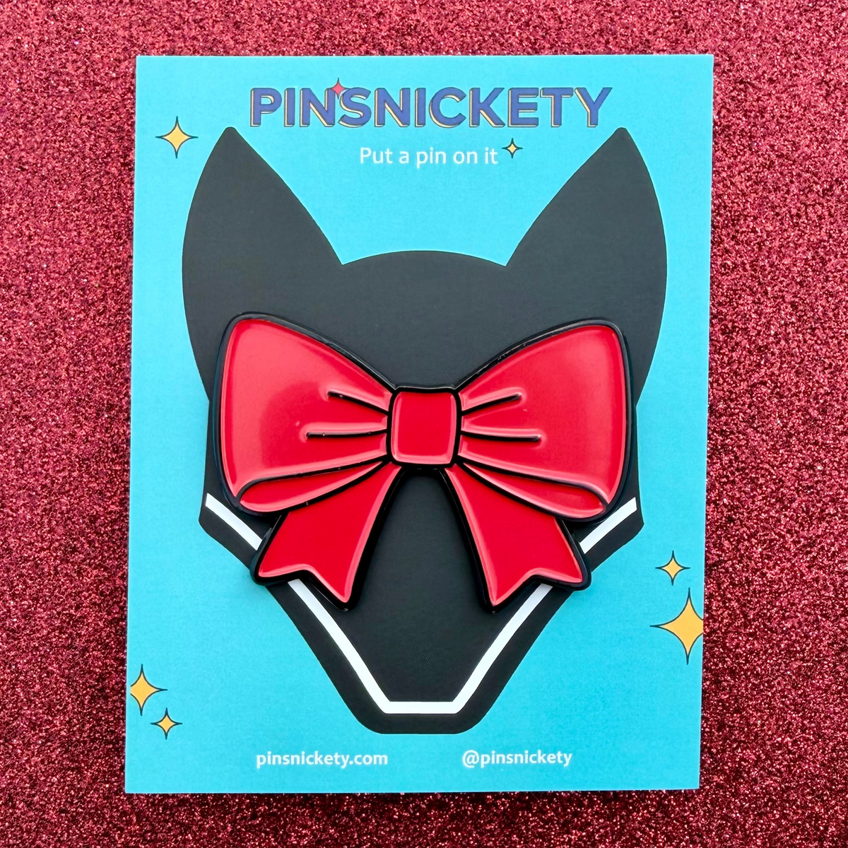 a pinsnickety back off red ribbon-shaped bonnet pin on its product packaging card in front of a red glitter background. the card is teal colored, has the brand name on it, and features an illustration of a horse's fly bonnet.