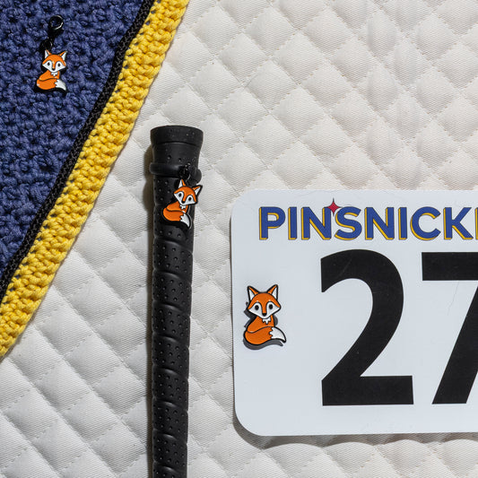 A saddle pad, bonnet and whip decorated with matching Fox Pinsnickety horse show number pins, a bridle charm and a crop charm.