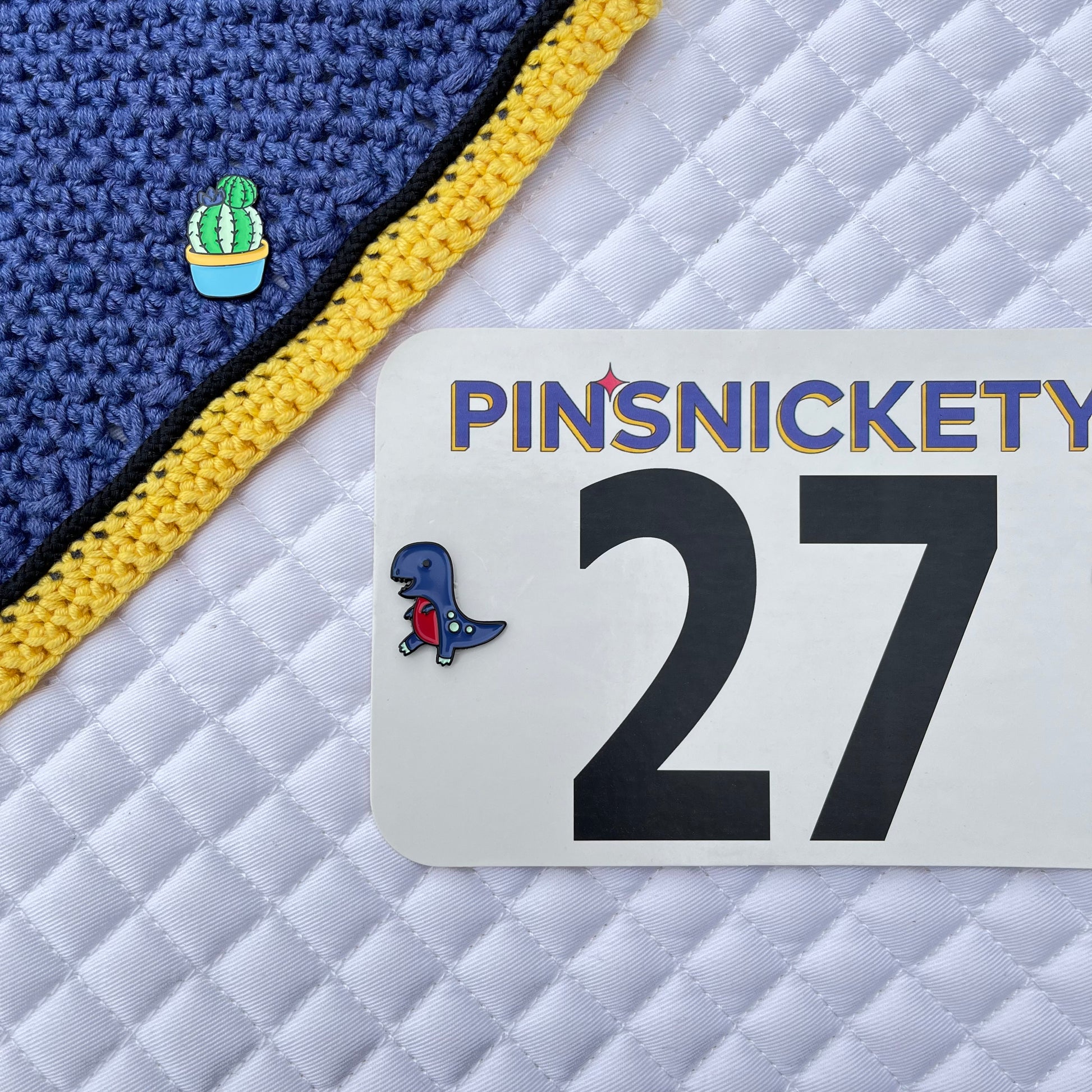 pinsnickety cactus pin on a fly bonnet and t-rex horse pin on a horse show number attached to a saddle pad