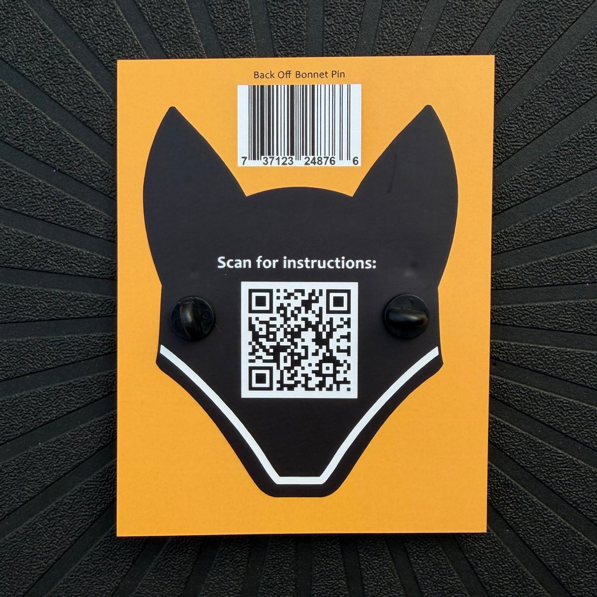 The yellow back of a Pinsnickety product packaging card with an illustration of a jumper fly bonnet, showing the rubber pin clasps of the back off red ribbon bonnet pin, a QR code, and a UPC code.