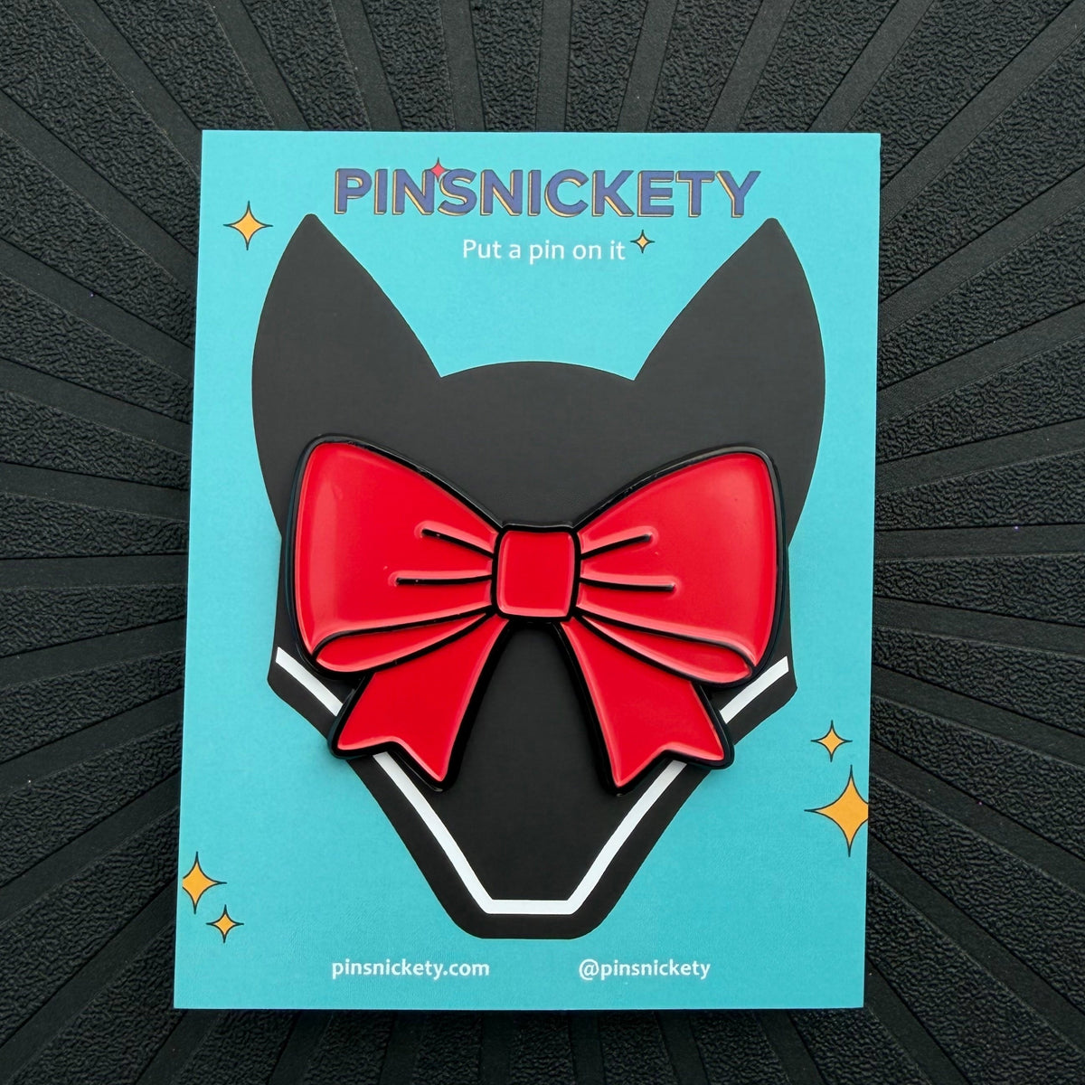 A back off red ribbon enamel bonnet pin displayed on a teal Pinsnickety card with an illustration of a jumper fly bonnet, set on a black textured background.