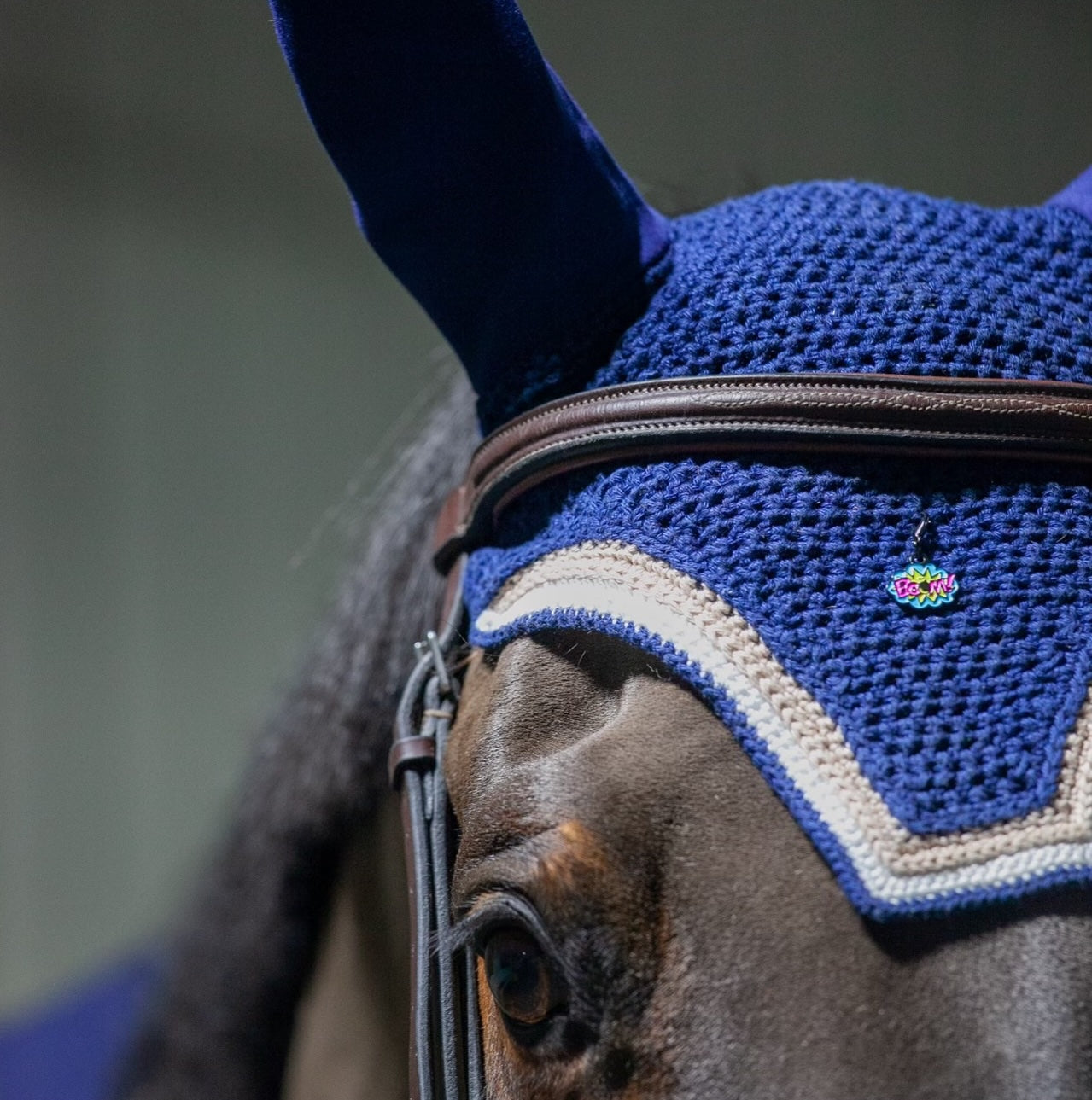 a closeup of a pinsnickety boom! bridle charm on a horse's blue fly bonnet