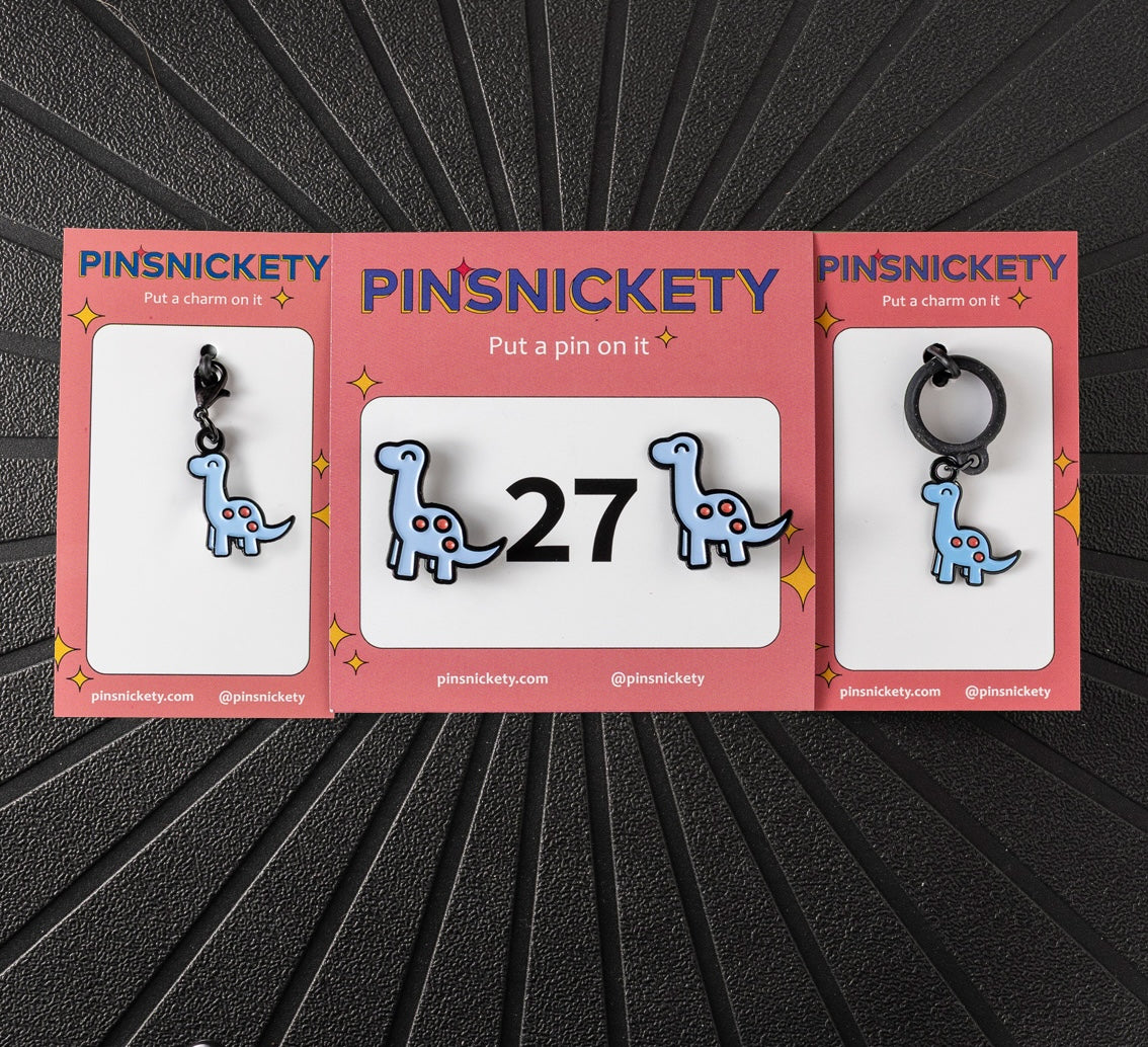 A matching triple combination set with Pinsnickety Brontosaurus horse show number pins, a bridle charm, and a crop charm in their product packaging on a black background.