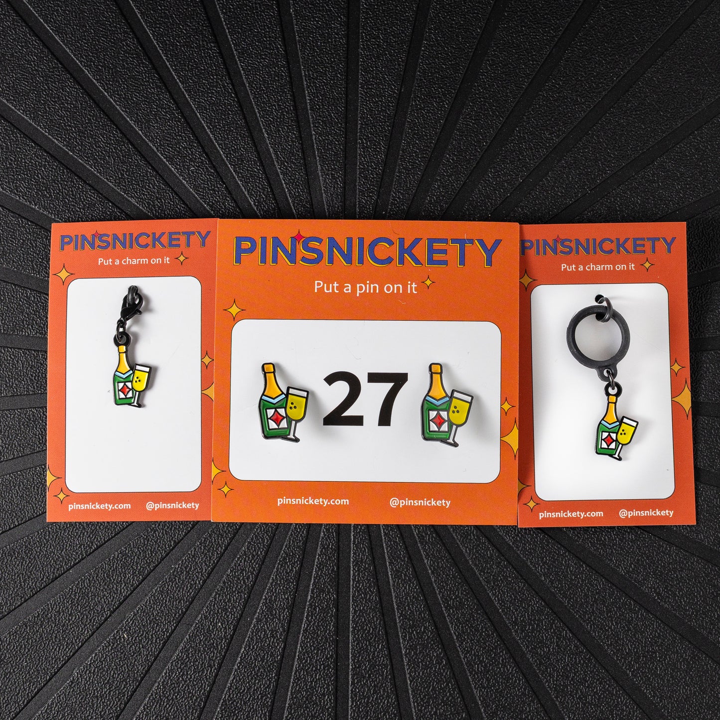 A matching triple combination set with Pinsnickety Champagne horse show number pins, a bridle charm, and a crop charm in their product packaging on a black background.