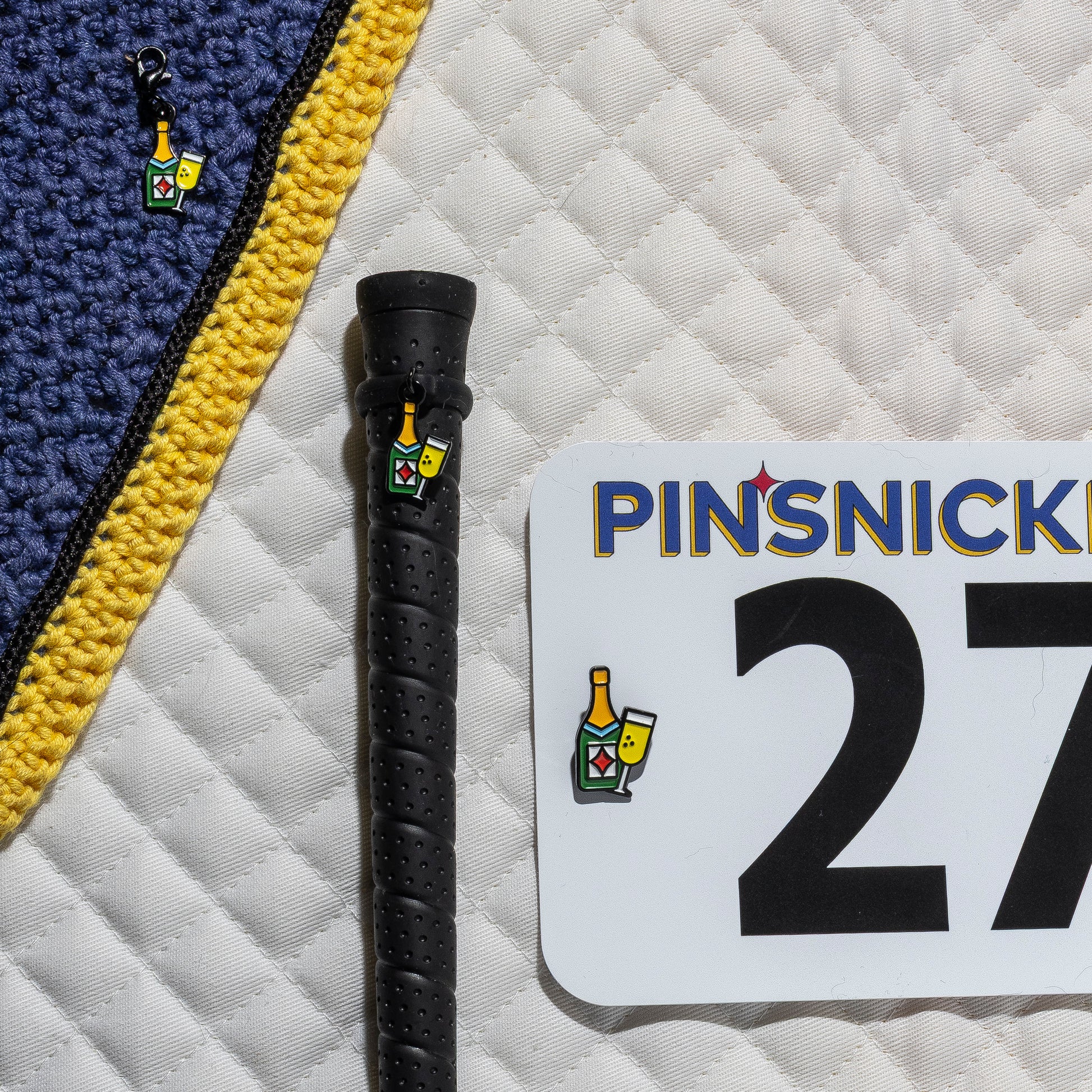 A saddle pad, bonnet and whip decorated with matching Champagne Pinsnickety horse show number pins, a bridle charm and a crop charm.