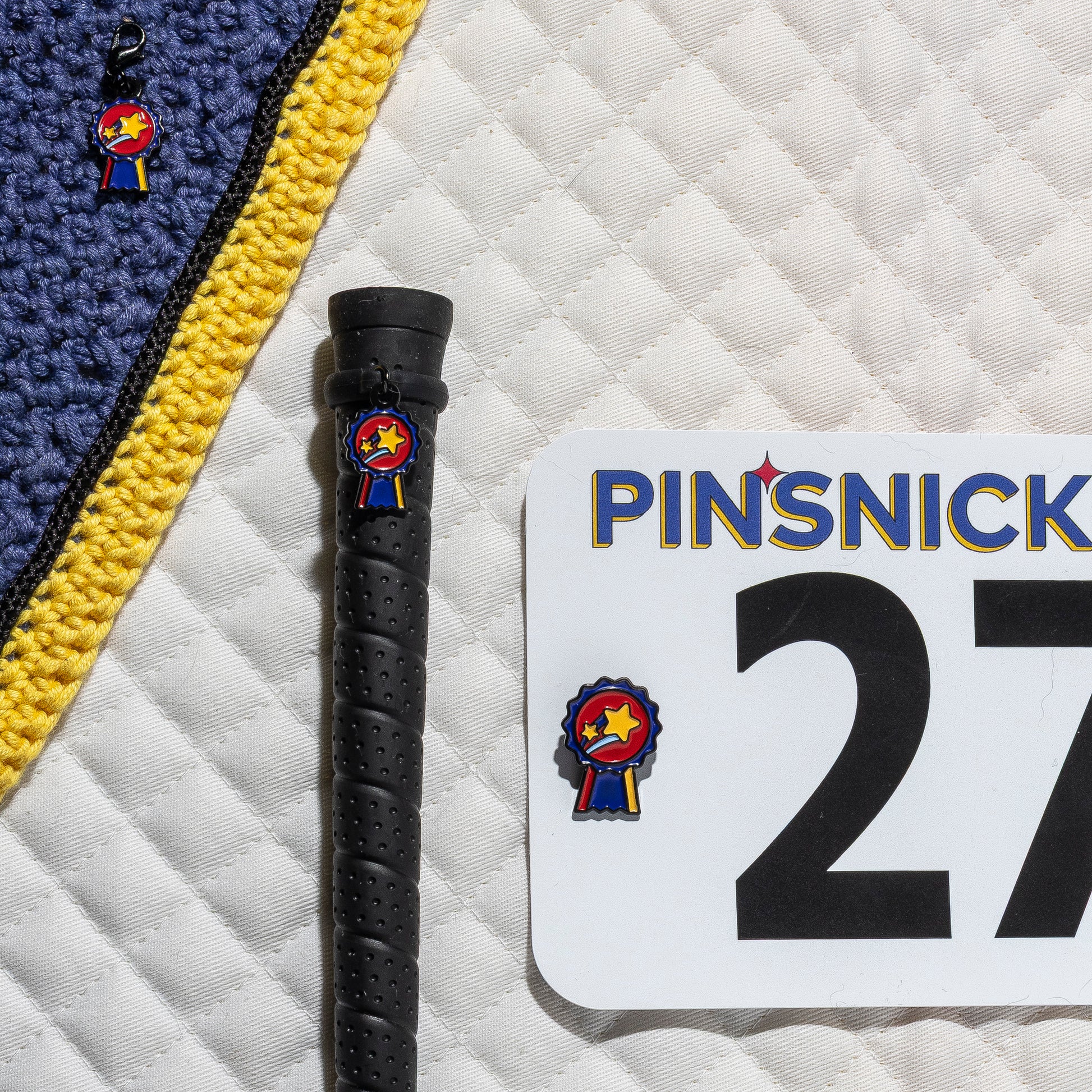 A saddle pad, bonnet and whip decorated with matching Champion Pinsnickety horse show number pins, a bridle charm and a crop charm.