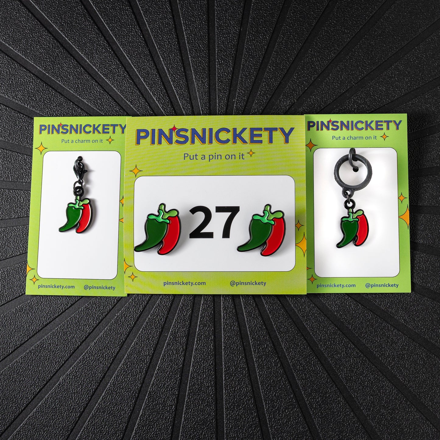 A matching triple combination set with Pinsnickety Chili Peppers horse show number pins, a bridle charm, and a crop charm in their product packaging on a black background.