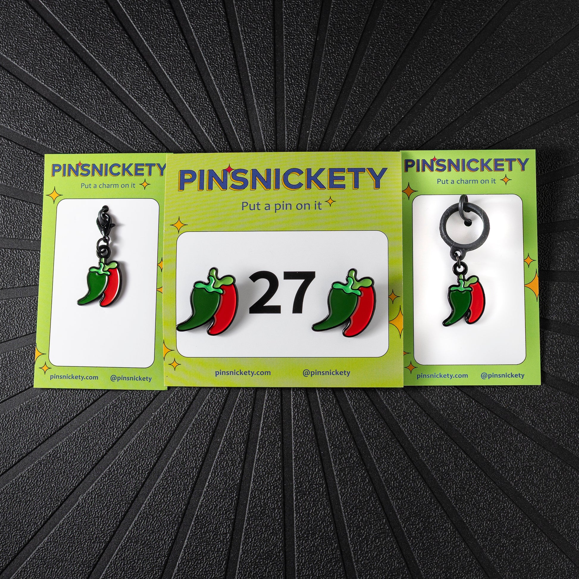 A matching triple combination set with Pinsnickety Chili Peppers horse show number pins, a bridle charm, and a crop charm in their product packaging on a black background.