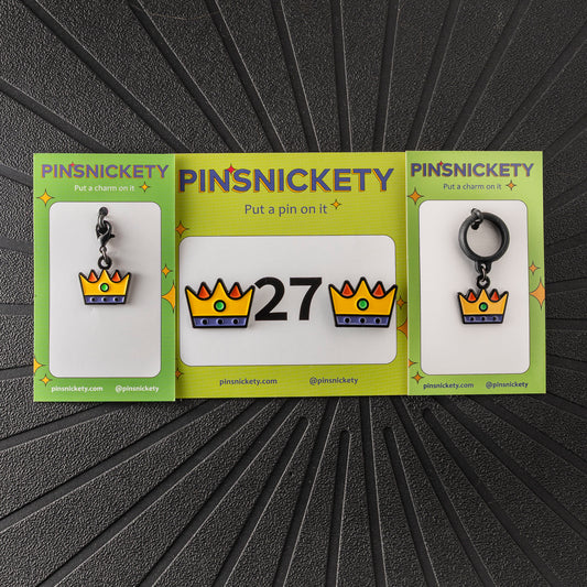 A matching triple combination set with Pinsnickety Crown horse show number pins, a bridle charm, and a crop charm in their product packaging on a black background.