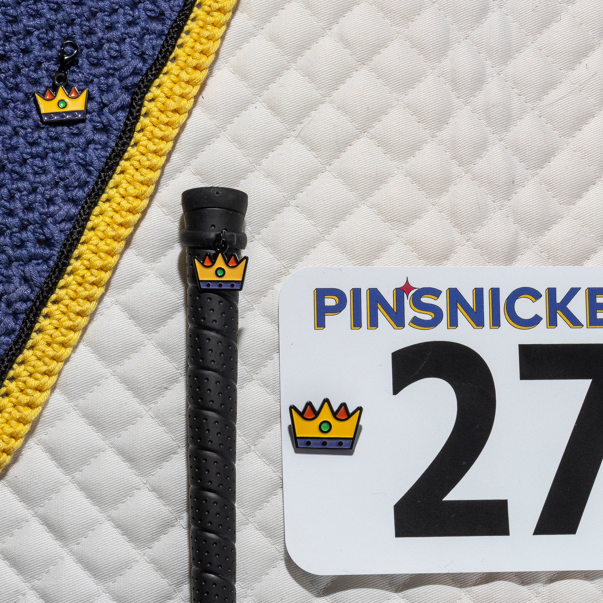 A saddle pad, bonnet and whip decorated with matching Crown Pinsnickety horse show number pins, a bridle charm and a crop charm.