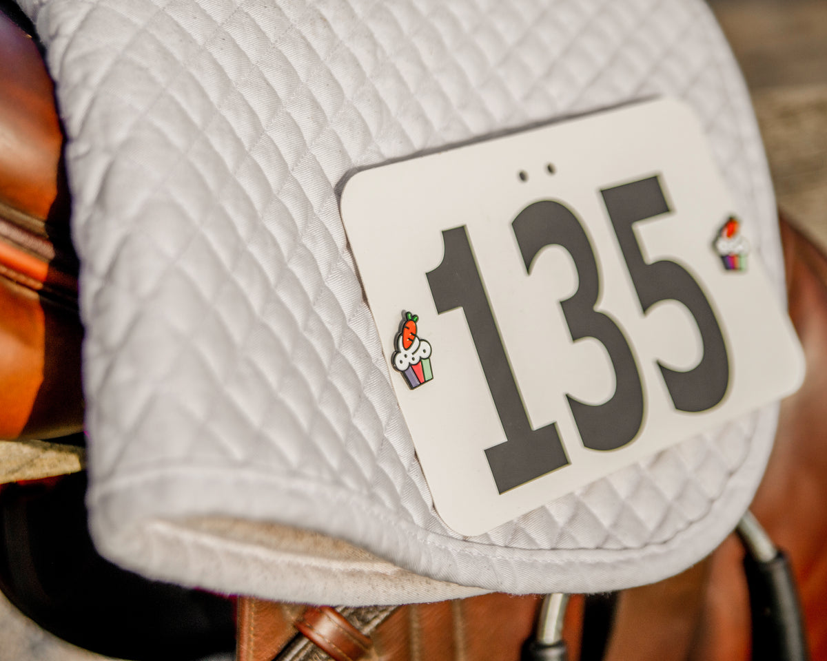 A close-up of a white quilted saddle pad with a white numbered attached with Pinsnickety Cupcake horse show number pins, placed on a brown leather saddle.