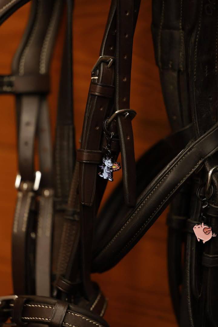 two bridles hang in a wood-walled tackroom. On each bridle is a pinsnickety bridle charm. The one on the left is a blue dragon clip charm, and the one on the right is a pink flying pig clip charm.