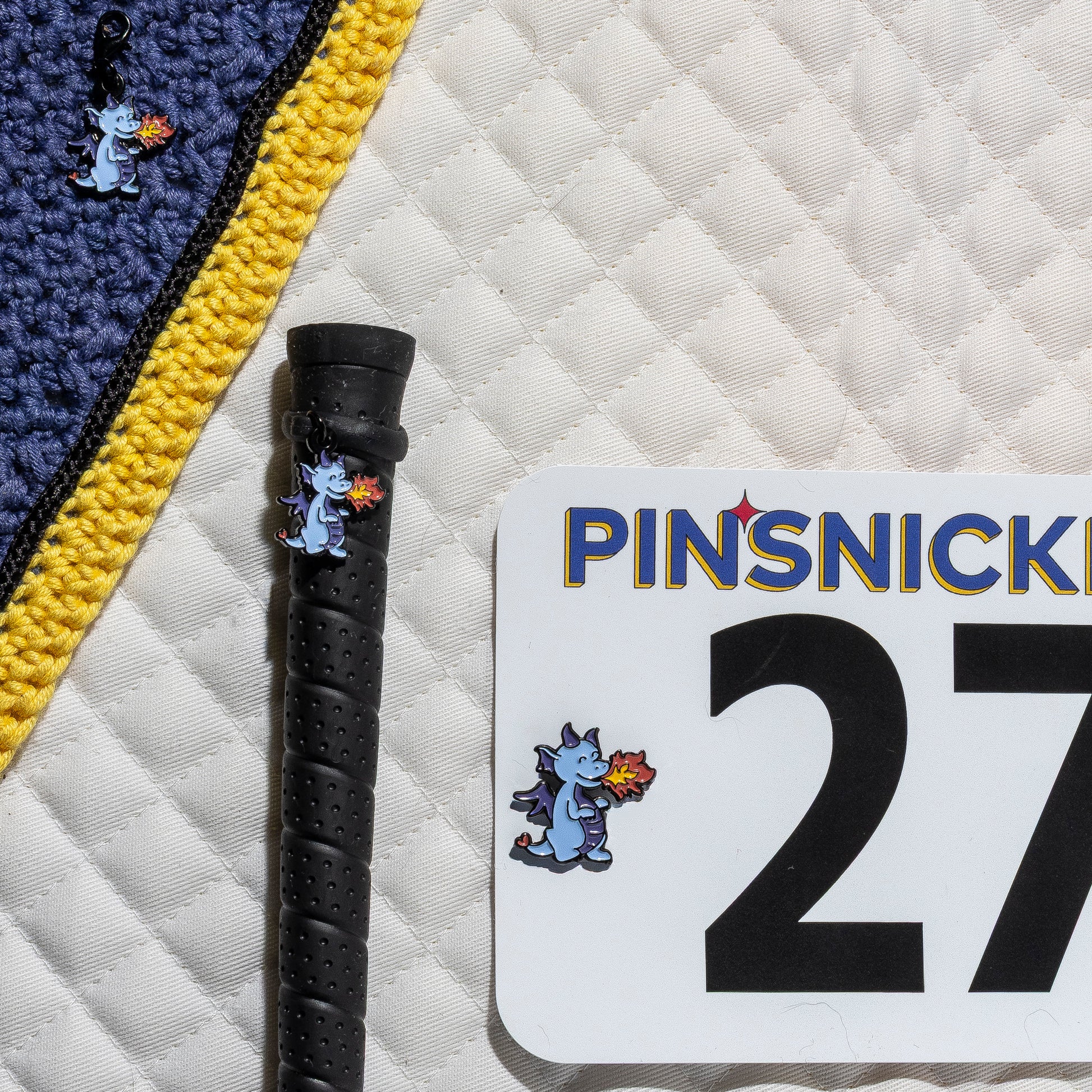 A saddle pad, bonnet and whip decorated with matching Dragon Pinsnickety horse show number pins, a bridle charm and a crop charm.