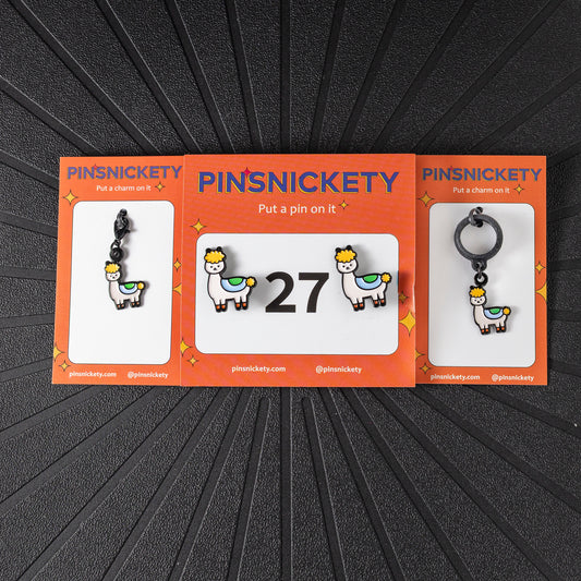 A matching triple combination set with Pinsnickety Drama llama horse show number pins, a bridle charm, and a crop charm in their product packaging on a black background.