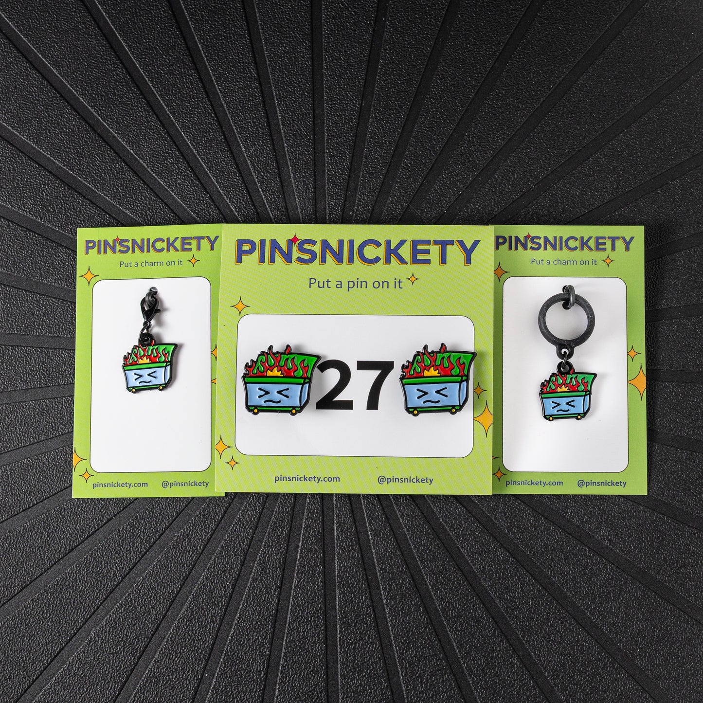 A matching triple combination set with Pinsnickety Dumpster Fire horse show number pins, a bridle charm, and a crop charm in their product packaging on a black background.