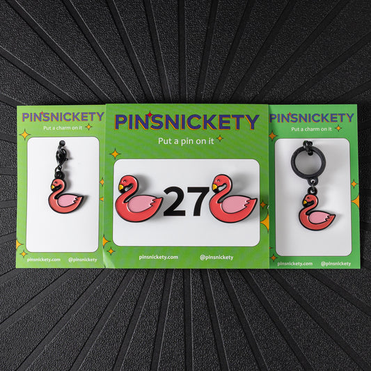 A matching triple combination set with Pinsnickety Flamingo horse show number pins, a bridle charm, and a crop charm in their product packaging on a black background.