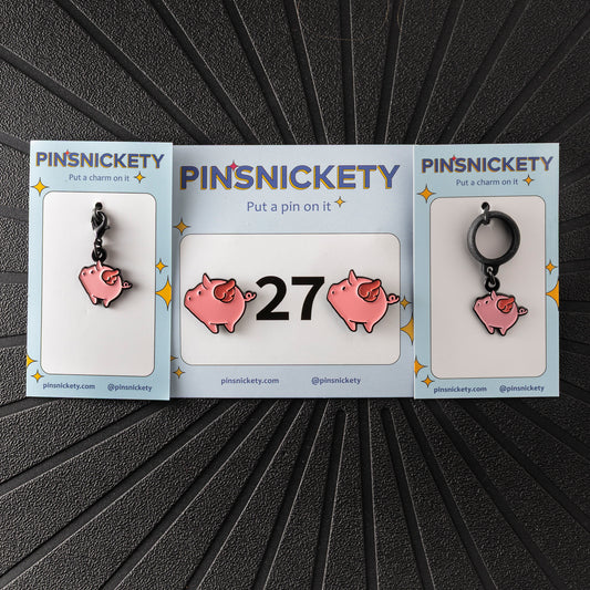A matching triple combination set with Pinsnickety Flying Pig horse show number pins, a bridle charm, and a crop charm in their product packaging on a black background.