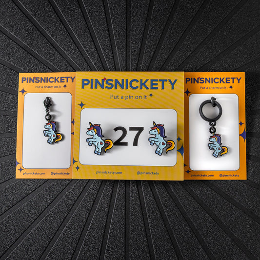 A matching triple combination set with Pinsnickety Flying Unicorn horse show number pins, a bridle charm, and a crop charm in their product packaging on a black background.
