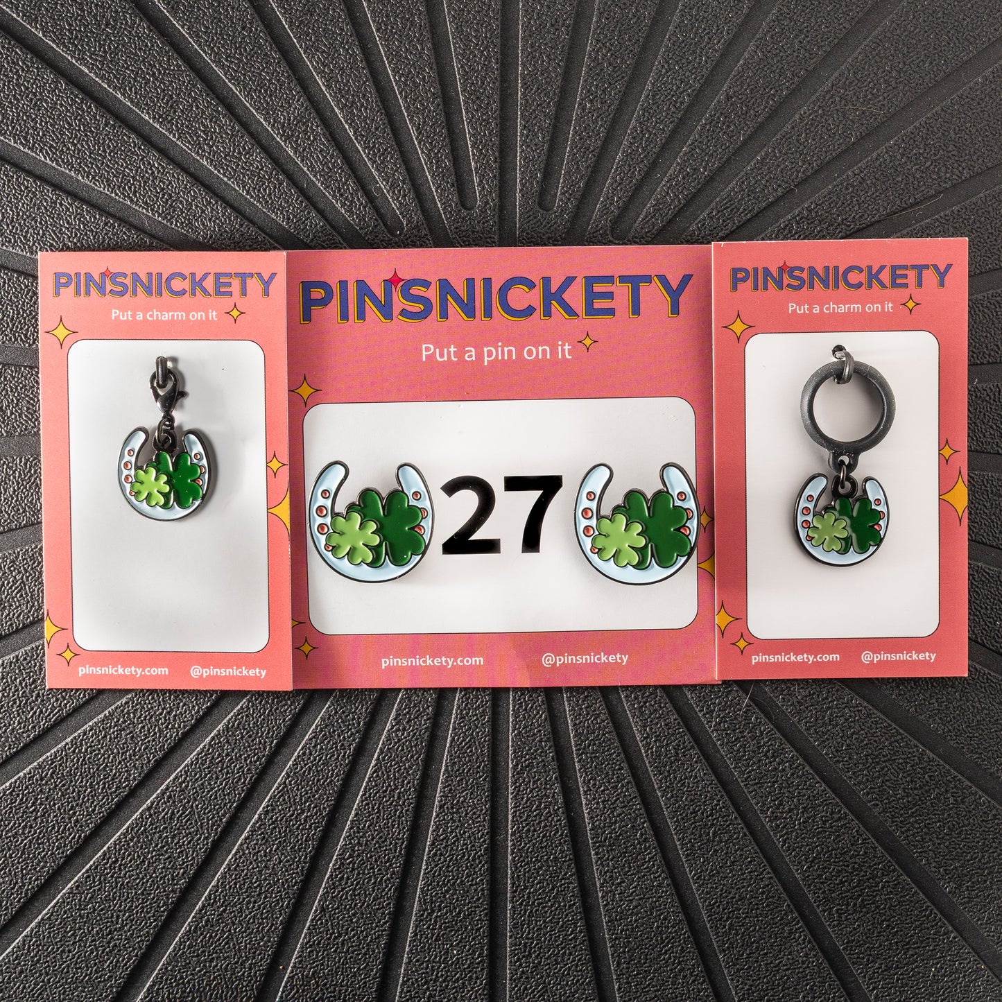 A matching triple combination set with Pinsnickety Horseshoe horse show number pins, a bridle charm, and a crop charm in their product packaging on a black background.