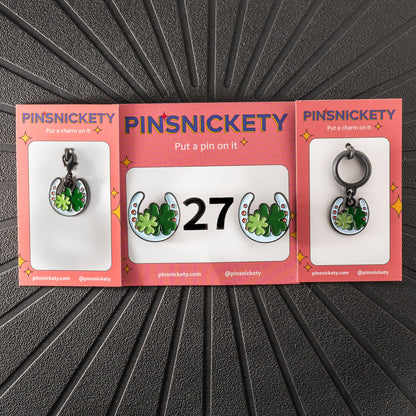 A matching triple combination set with Pinsnickety Horseshoe horse show number pins, a bridle charm, and a crop charm in their product packaging on a black background.