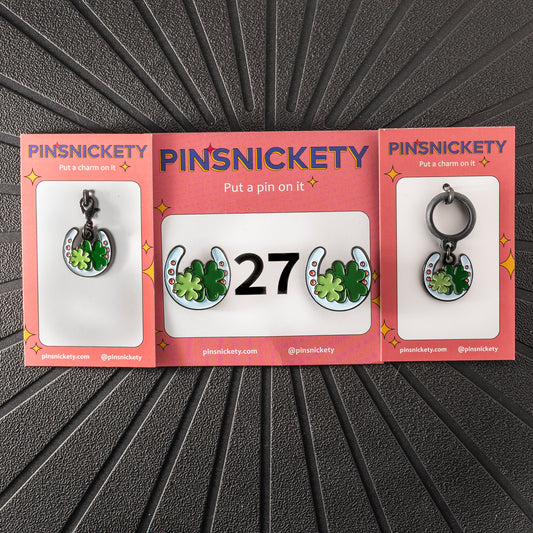 A matching triple combination set with Pinsnickety Horseshoe horse show number pins, a bridle charm, and a crop charm in their product packaging on a black background.