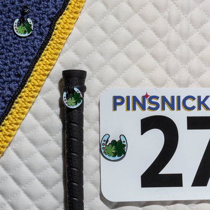A saddle pad, bonnet and whip decorated with matching Hoeseshoe Pinsnickety horse show number pins, a bridle charm and a crop charm.