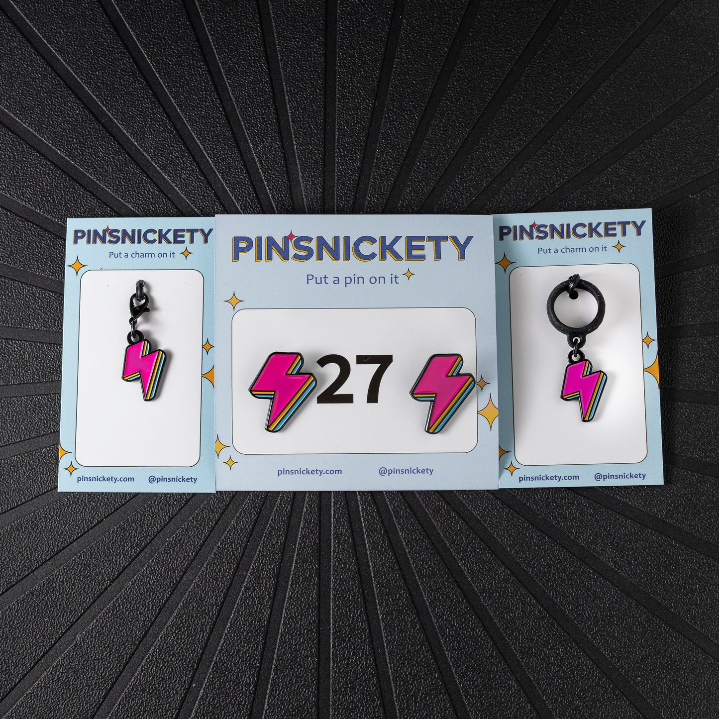 A matching triple combination set with Pinsnickety Lightning Bolt horse show number pins, a bridle charm, and a crop charm in their product packaging on a black background.