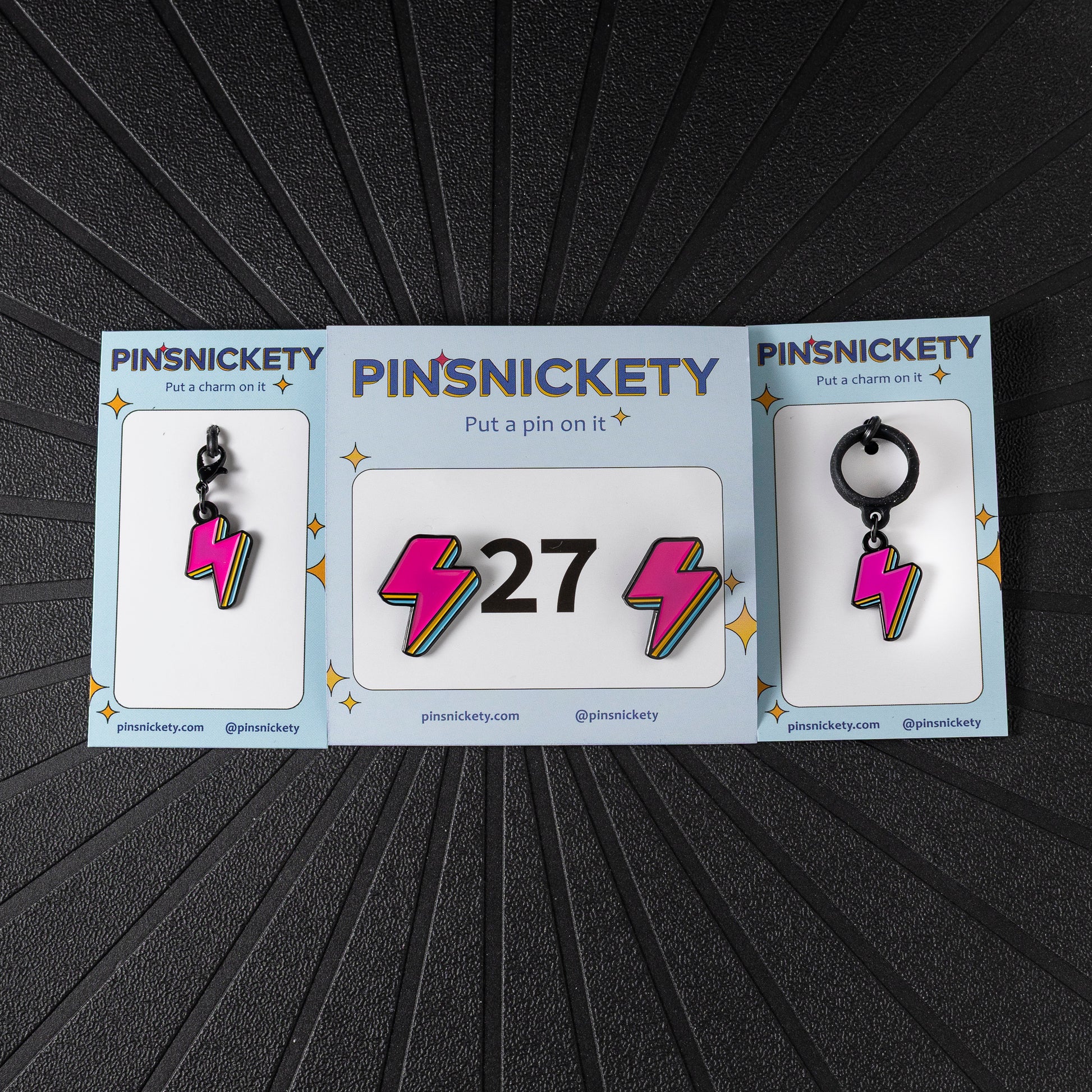 A matching triple combination set with Pinsnickety Lightning Bolt horse show number pins, a bridle charm, and a crop charm in their product packaging on a black background.