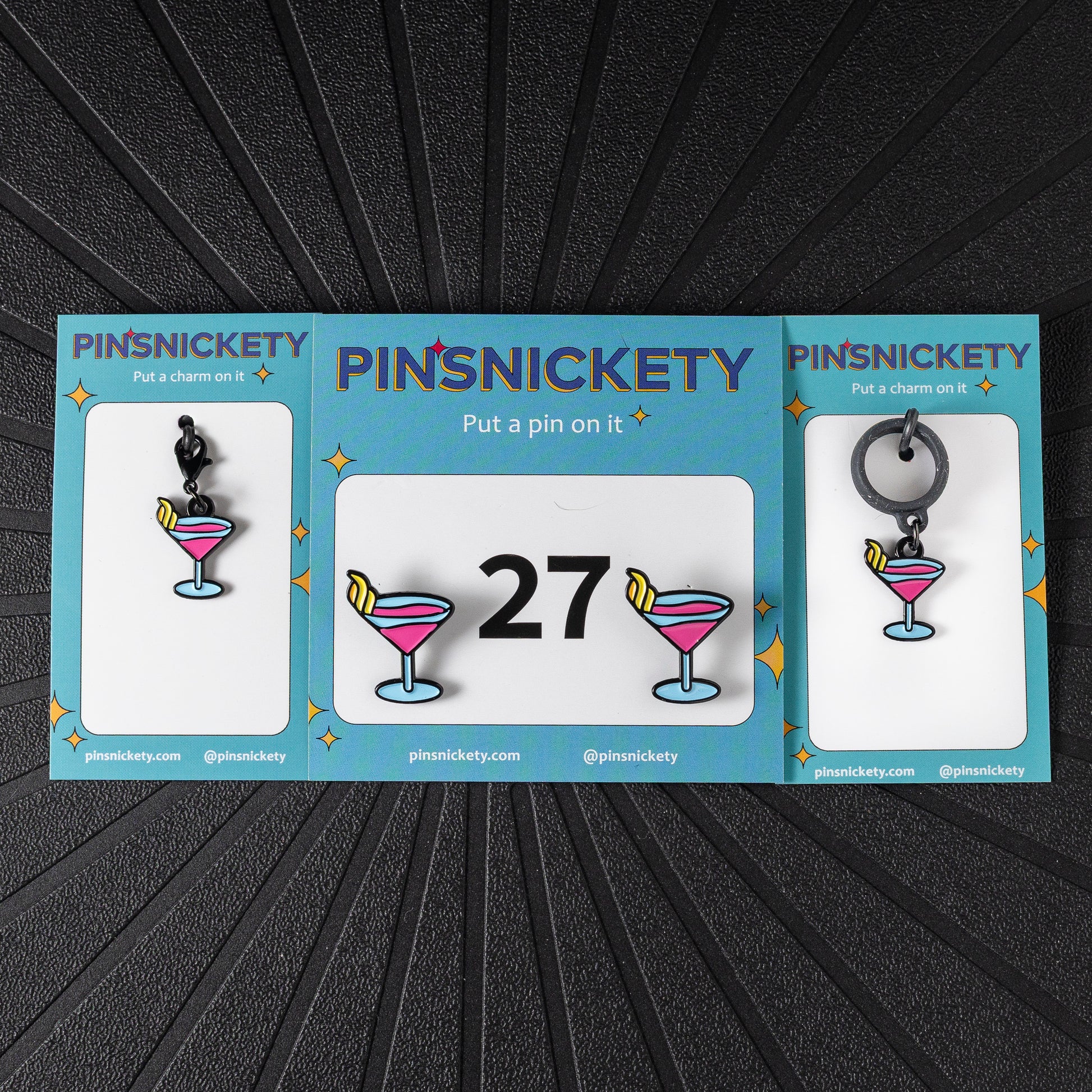 A matching triple combination set with Pinsnickety Martini horse show number pins, a bridle charm, and a crop charm in their product packaging on a black background.