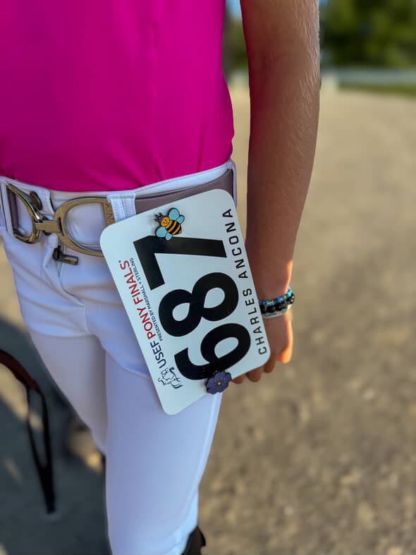 a close up of a pony jumper rider at pony finals waiting for the jog. She is wearing a bright pink shirt and white breeches. Her show number is attached to her belt loop with a Pinsnickety Queen Bee horse show number pin.