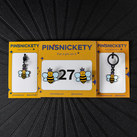 A matching triple combination set with Pinsnickety Queen Bee horse show number pins, a bridle charm, and a crop charm in their product packaging on a black background.