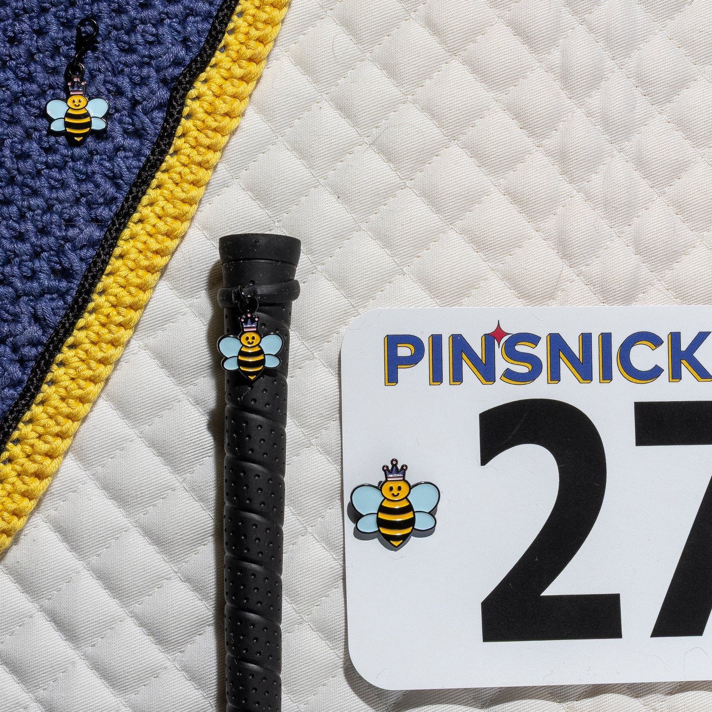 A saddle pad, bonnet and whip decorated with matching Queen Bee Pinsnickety horse show number pins, a bridle charm and a crop charm.