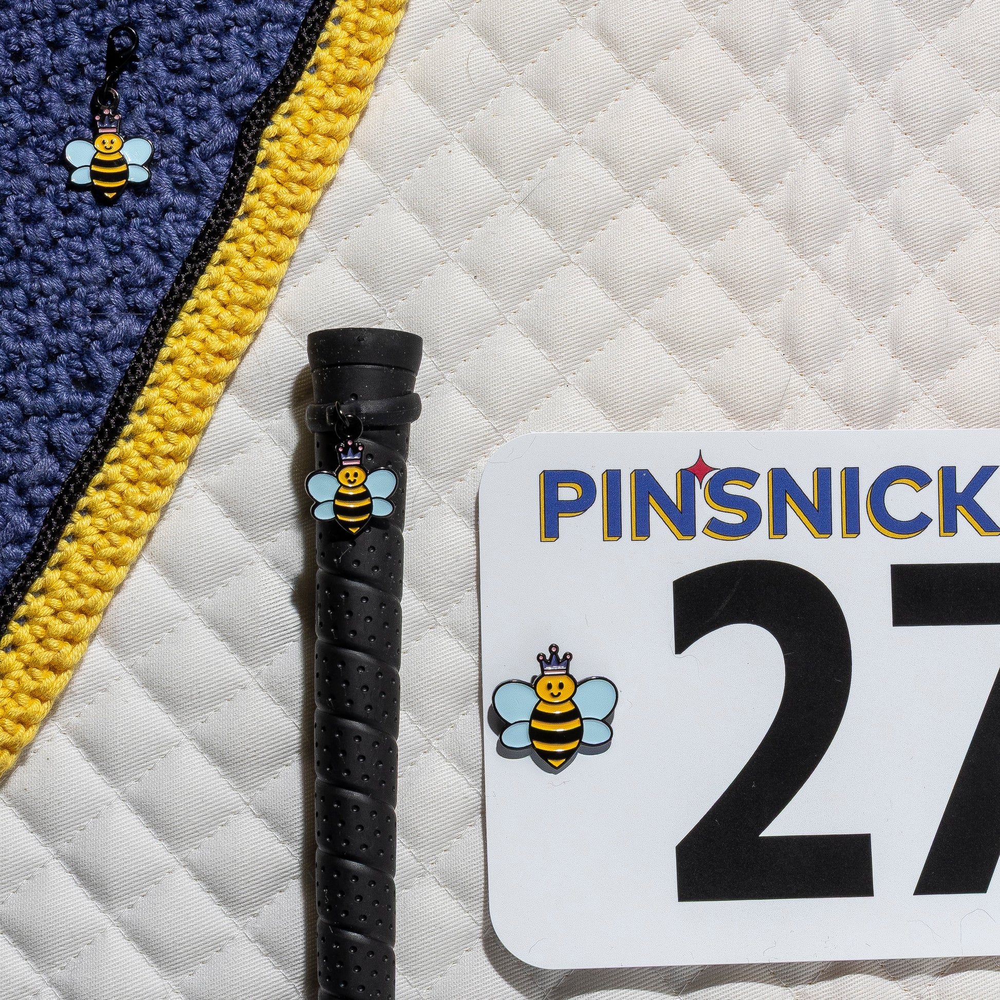 A saddle pad, bonnet and whip decorated with matching Queen Bee Pinsnickety horse show number pins, a bridle charm and a crop charm.