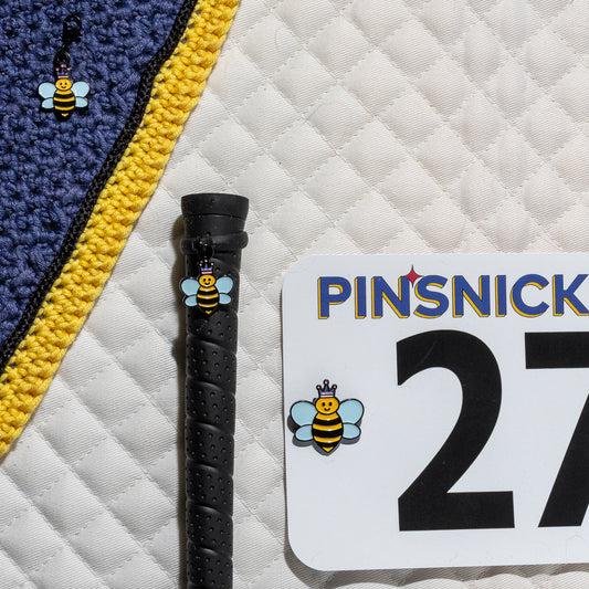 A saddle pad, bonnet and whip decorated with matching Queen Bee Pinsnickety horse show number pins, a bridle charm and a crop charm.
