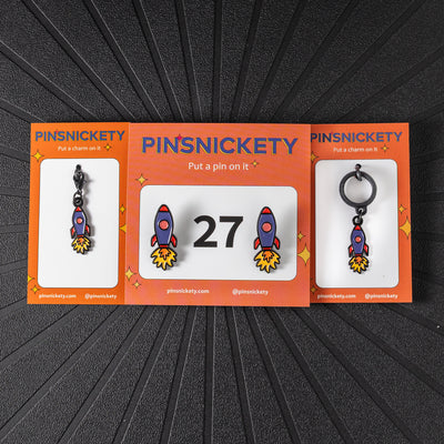 A matching triple combination set with Pinsnickety Rocket Ship horse show number pins, a bridle charm, and a crop charm in their product packaging on a black background.
