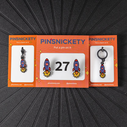 A matching triple combination set with Pinsnickety Rocket Ship horse show number pins, a bridle charm, and a crop charm in their product packaging on a black background.