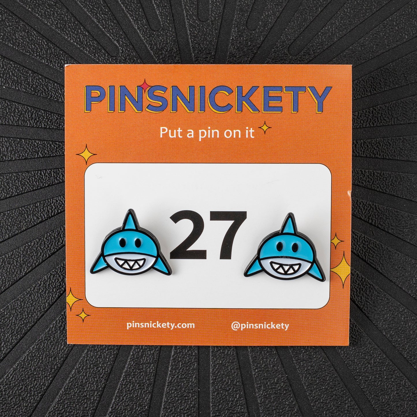 A pair of pinsnickety horse show number pins featuring blue sharks on either side of the number 27, presented on its orange backing card against a dark textured background.