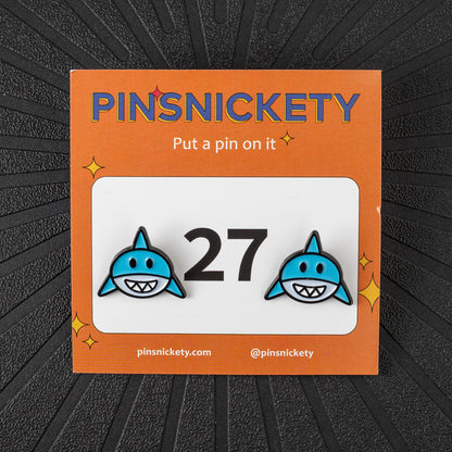 A pair of pinsnickety horse show number pins featuring blue sharks on either side of the number 27, presented on its orange backing card against a dark textured background.