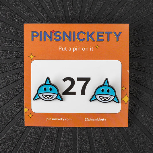 A pair of pinsnickety horse show number pins featuring blue sharks on either side of the number 27, presented on its orange backing card against a dark textured background.