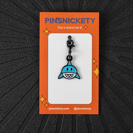 A blue shark bridle charm with a black clasp, displayed on an orange backing card labeled PINSNICKETY, set against a dark textured background.